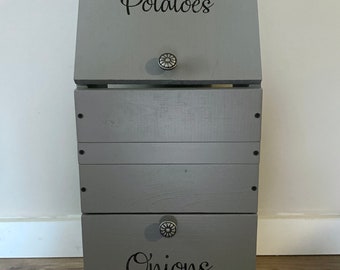 May include: A gray wooden storage bin with two compartments. The top compartment has a hinged lid and is labeled "Potatoes". The bottom compartment is labeled "Onions".