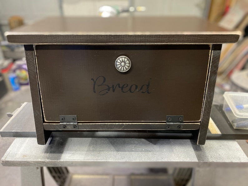 K&ouml;nnte beinhalten: Eine dunkelbraune Brotbox aus Holz mit einem Scharnierdeckel und einem silbernen Knopf. Der Deckel tr&auml;gt eine schwarze Aufschrift mit der Aufschrift "Brot".