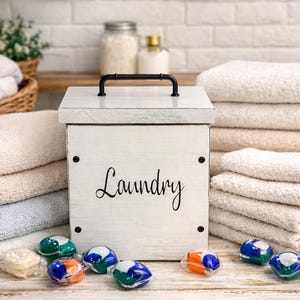 May include: A white wooden laundry box with a black handle and the word "Laundry" in script. Stacks of folded towels in various shades of white and beige surround the box. Laundry pods in blue, green, and orange are scattered in front.