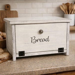 May include: A white wooden bread box with a distressed finish. The word "Bread" is written in a script font on the front. The box has a knob and black hinges. A loaf of bread and a cutting board are in the foreground.
