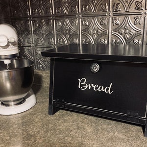K&ouml;nnte beinhalten: Schwarze Brotbox aus Holz mit einer wei&szlig;en Aufschrift "Brot". Die Box hat einen Metallknopf und Scharniere. Die Box steht auf einer Arbeitsplatte neben einem wei&szlig;en Standmixer.