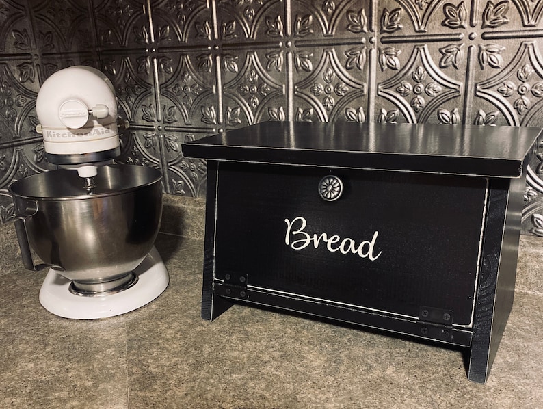 K&ouml;nnte beinhalten: Schwarze Brotbox aus Holz mit einer wei&szlig;en Aufschrift "Brot". Die Box hat einen Metallknopf und Scharniere. Die Box steht auf einer Arbeitsplatte neben einem wei&szlig;en Standmixer.