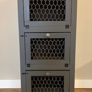 May include: A gray wooden cabinet with three drawers. Each drawer has a wire mesh front and a metal knob. The cabinet is standing on a wood floor.