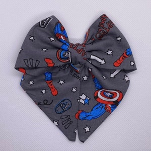 Puede incluir: Un lazo de tela gris con temática de Capitán América. El lazo está decorado con estrellas blancas, imágenes de Capitán América en rojo y azul, y el texto "Capitán América".