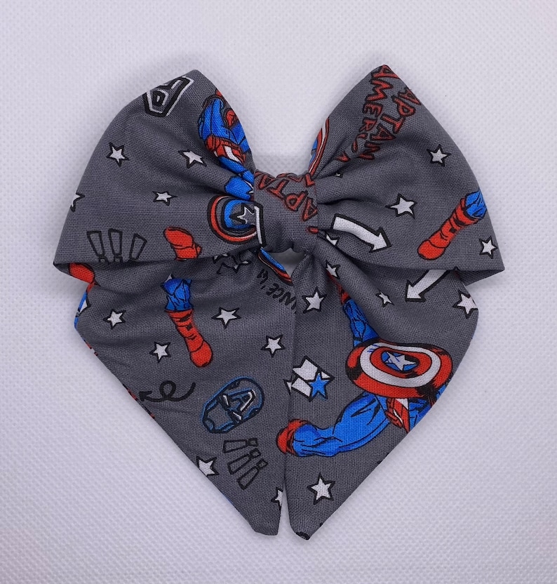 May include: A gray fabric bow with a Captain America theme. The bow is decorated with white stars, red and blue Captain America images, and the text "Captain America".