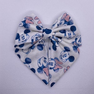 May include: A white fabric bow with a blue and pink Mickey Mouse print. The bow is tied in a classic shape with a center loop.