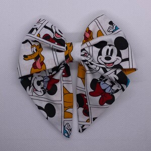 May include: A white bow with a cartoon print of Mickey Mouse, Minnie Mouse, and Pluto. The bow is made of fabric and has a white background with black outlines.