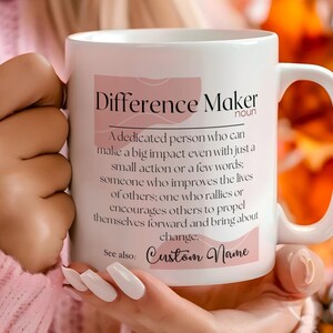 May include: White ceramic mug with a pink and white design. The mug features the text "Difference Maker" and a definition of the word. The text "Custom Name" appears at the bottom of the mug.