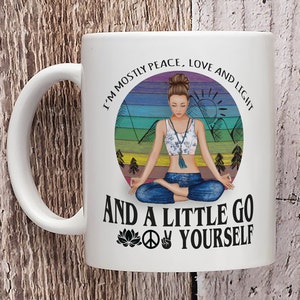 May include: White ceramic mug with a colorful illustration of a woman meditating in a forest setting. The text reads "I'm mostly peace, love and light and a little go yourself" with a peace sign and a lotus flower.