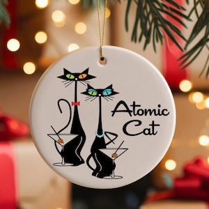 May include: A round, white ceramic ornament featuring two stylized black cats with martini glasses. The cats have colorful eyes and are adorned with a bow tie and necklace. The text "Atomic Cat" is written in a script font.