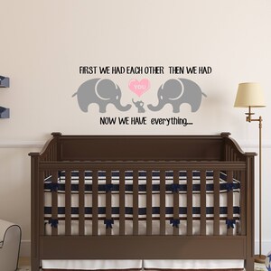 Pu&ograve; includere: Un adesivo da parete per l'asilo nido con due elefanti grigi e un elefante pi&ugrave; piccolo al centro. Gli elefanti tengono un cuore rosa con la parola "you" all'interno. Il testo dice "First we had each other, then we had you. Now we have everything..."