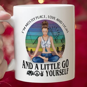 May include: White ceramic mug with a colorful illustration of a woman meditating in a mountain landscape. The text reads "I'm mostly peace, love and light and a little go yourself" with a peace sign and a lotus flower.