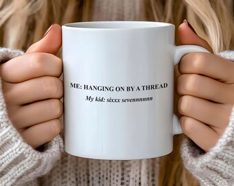 May include: White ceramic mug with the text "ME: HANGING ON BY A THREAD" and "My kid: sixxx sevennnnnnn" in black. The mug is held by a person wearing a cream-colored sweater.
