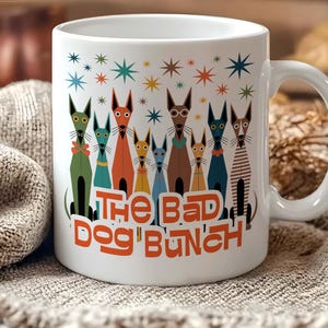 May include: White ceramic mug featuring a colorful illustration of stylized dogs with long snouts. The dogs are in various colors and wear accessories. The text "THE BAD DOG BUNCH" is printed in orange. The mug has a white handle.