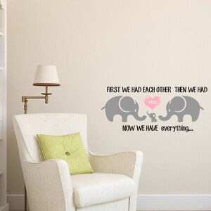 Pu&ograve; includere: Un muro grigio con un adesivo in vinile di due elefanti e un elefante bambino con un cuore che dice "you". Il testo dice "First we had each other, then we had you. Now we have everything..."