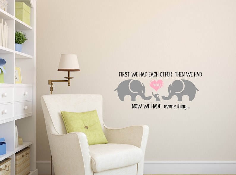 Pu&ograve; includere: Un muro grigio con un adesivo in vinile di due elefanti e un elefante bambino con un cuore che dice "you". Il testo dice "First we had each other, then we had you. Now we have everything..."