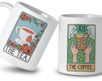 May include: Two white ceramic mugs with illustrations of tarot cards. The first mug features a teacup with a teabag and the text "THE TEA." The second mug features a hand holding a cup of coffee with a straw and the text "XII THE COFFEE."