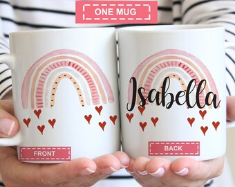 May include: White ceramic mug with a pink and orange watercolor rainbow design and red hearts. The back of the mug has the name "Isabella" written in cursive.