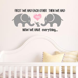 Pu&ograve; includere: Un adesivo da parete per l'asilo con due elefanti grigi e un cuore rosa che dice "You". Gli elefanti si guardano e il cuore &egrave; tra loro. Il testo dice "First we had each other then we had you now we have everything..."