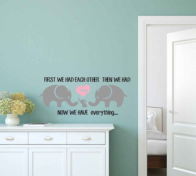 Pu&ograve; includere: Un adesivo da parete con due elefanti grigi e un cuore rosa che dice "You". Il testo dice "First we had each other, then we had you. Now we have everything..."