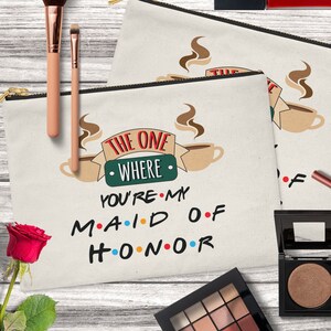 May include: A beige canvas makeup bag with a zipper closure. The bag features a graphic of two coffee mugs with steam rising from them. The text "The One Where You're My Maid of Honor" is printed on the bag in a playful font.