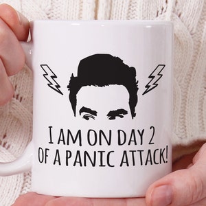 Può includere: Una tazza in ceramica bianca con una silhouette nera di un viso maschile con fulmini sopra la testa. Il testo sulla tazza recita "I am on day 2 of a panic attack!"