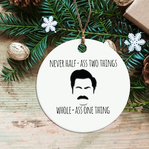 May include: White round Christmas ornament with a black and white illustration of Ron Swanson from the TV show Parks and Recreation. The ornament has the quote "Never half-ass two things whole-ass one thing".