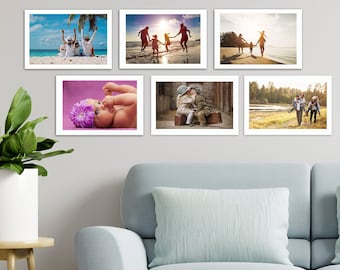 May include: Six framed photographs with white borders are displayed on a gray wall. The photos depict family moments: people on a beach, a baby with a flower, and children playing. A green plant in a white pot and a blue sofa are in the foreground.