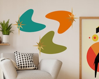 Puede incluir: Tres pegatinas de pared en forma de boomerang de estilo moderno de mediados de siglo en verde azulado, naranja y verde oliva con acentos de estrella dorada. Las pegatinas están en una pared blanca con un sillón gris y una impresión enmarcada en el fondo.