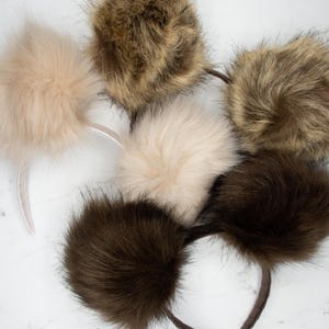 May include: A selection of faux fur ear muffs in shades of brown and beige. The ear muffs are attached to headbands. The fur appears soft and fluffy, with a variety of textures and colours, including light beige, dark brown, and mixed tones.