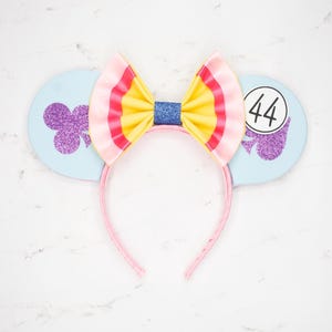May include: A light blue headband with mouse ears, a colorful bow, and a pink headband. The ears have a purple glitter design and the number 44 in a circle. The bow has pink, yellow, and blue stripes.