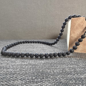Peut inclure: Un collier de perles en pierre de lave noire avec un fermoir argenté. Le collier est posé sur une surface texturée grise.