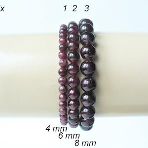 Peut inclure: Quatre bracelets de perles de grenat en différentes tailles, étiquetés 4 mm, 6 mm et 8 mm. Les bracelets sont sur une surface brun clair.