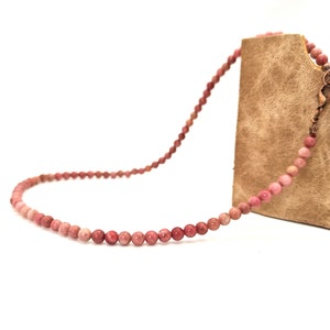 May include: A pink beaded necklace with a copper clasp. The necklace is draped over a brown leather bag.
