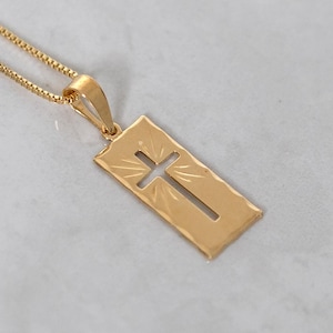 May include: A gold-colored pendant necklace featuring a rectangular pendant with a cut-out cross design. The pendant hangs from a delicate gold chain. The necklace is displayed on a light gray surface, highlighting the jewelry's details.