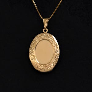 May include: A gold-toned oval locket pendant suspended from a matching thin box chain. The locket features a smooth, polished centre panel surrounded by an ornate, raised floral border with engraved leaf and flower details throughout the frame.