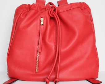 May include: A bright red leather backpack with a drawstring closure and a gold zipper. The backpack has a casual, relaxed style.