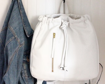 May include: White leather backpack with a drawstring closure and a gold zipper. The backpack is hanging on a hook.