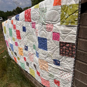 May include: A patchwork quilt featuring a grid of white squares interspersed with smaller, colorful patterned squares in shades of red, blue, green, yellow, and pink. The quilt has a dark gray floral border and curved, decorative machine quilting.
