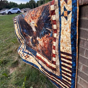 May include: A quilted fabric throw featuring a central graphic of a bald eagle with wings spread against a fiery orange and brown background. The border incorporates red and white stripes, blue sections with star patterns, and text reading 1776 and USA.