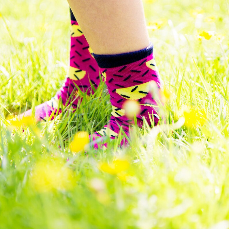 May include: A pair of pink socks with yellow and black geometric patterns. The socks are worn by a person standing in a grassy field.