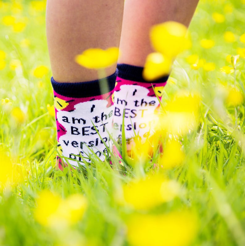 May include: A pair of colorful socks with the text "I am the best version of me" and "I'm the best version of me" on them. The socks are worn by a person standing in a field of yellow flowers.
