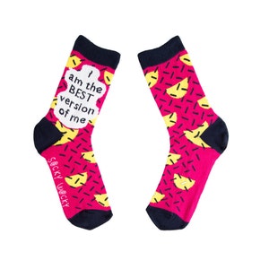 May include: A pair of pink socks with a yellow and black geometric pattern. The socks have the text "I am the BEST version of me" on one sock.