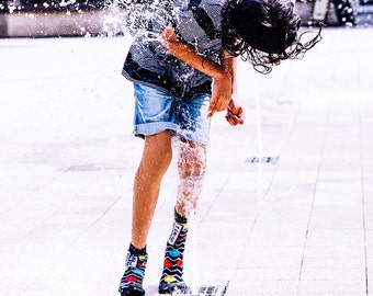 Könnte beinhalten: Eine Person wird von einem Springbrunnen durchnässt, wobei Wasser über Kopf und Körper fließt. Sie trägt ein gestreiftes Hemd, Jeansshorts und bunte Socken. Die Szene fängt einen Moment spielerischen Spaßes an einem sonnigen Tag ein.