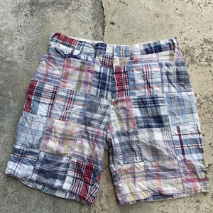 May include: Patchwork plaid shorts in shades of blue, red, and white. The shorts feature a button closure and pockets. The fabric appears to be a lightweight cotton, suitable for warm weather.