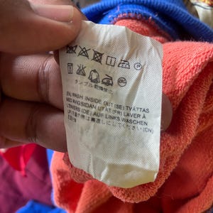 K&ouml;nnte beinhalten: Ein wei&szlig;es Pflegeetikett an einem korallenfarbenen Kleidungsst&uuml;ck mit blauem Rippkragen. Das Etikett zeigt schwarze Pflegesymbole und gedruckte Anweisungen in mehreren Sprachen, darunter Englisch, Schwedisch, Franz&ouml;sisch, Deutsch und Japanisch.