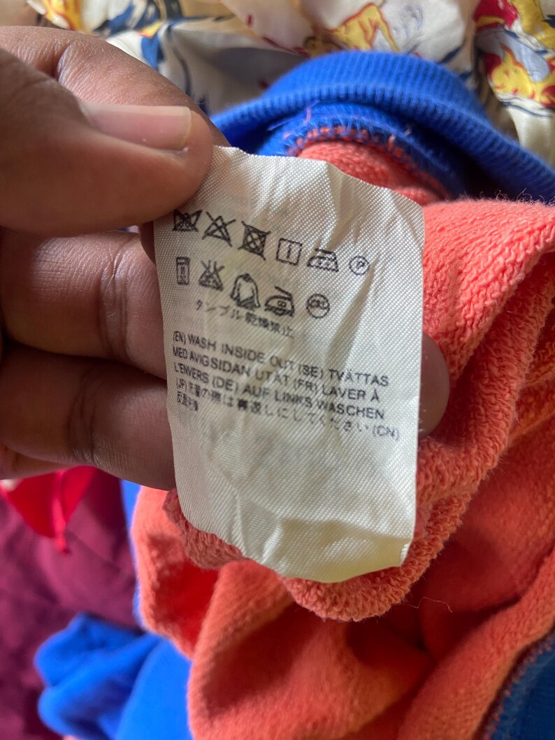 K&ouml;nnte beinhalten: Ein wei&szlig;es Pflegeetikett an einem korallenfarbenen Kleidungsst&uuml;ck mit blauem Rippkragen. Das Etikett zeigt schwarze Pflegesymbole und gedruckte Anweisungen in mehreren Sprachen, darunter Englisch, Schwedisch, Franz&ouml;sisch, Deutsch und Japanisch.