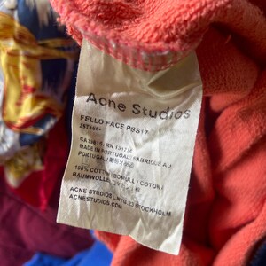 K&ouml;nnte beinhalten: Ein wei&szlig;es Textiletikett an einem korallenfarbenen Kleidungsst&uuml;ck aus Baumwolle. Das Etikett enth&auml;lt den Text Acne Studios, FELLO FACE PSS17, 25T166-, CA39815, RN 131738, MADE IN PORTUGAL, 100% COTTON und ACNE STUDIOS L.NYG.23 STOCKHOLM ACNESTUDIOS.COM.