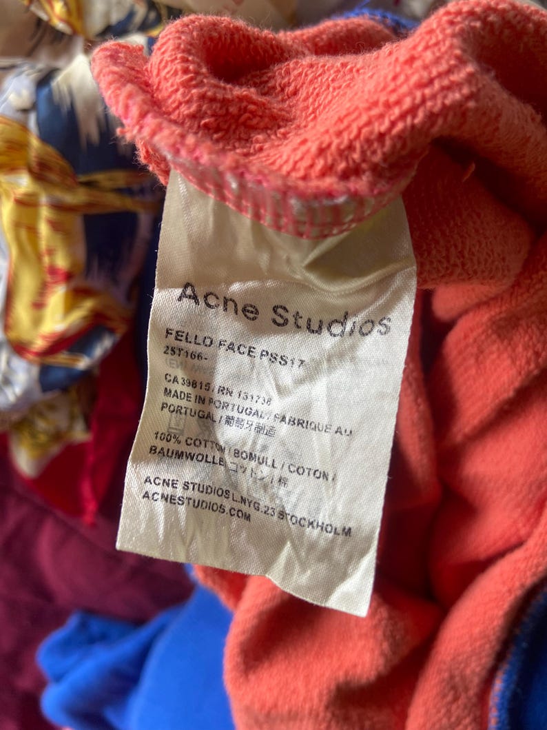 K&ouml;nnte beinhalten: Ein wei&szlig;es Textiletikett an einem korallenfarbenen Kleidungsst&uuml;ck aus Baumwolle. Das Etikett enth&auml;lt den Text Acne Studios, FELLO FACE PSS17, 25T166-, CA39815, RN 131738, MADE IN PORTUGAL, 100% COTTON und ACNE STUDIOS L.NYG.23 STOCKHOLM ACNESTUDIOS.COM.