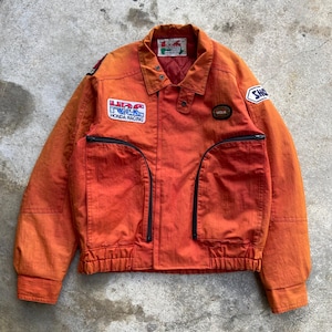May include: An orange jacket with a faded aesthetic, featuring a collar, long sleeves, and a ribbed hem. The front displays a Honda Racing HRC patch, a small oval badge, and two large zippered pockets. A SHO patch is on the sleeve and a label is at the neck.